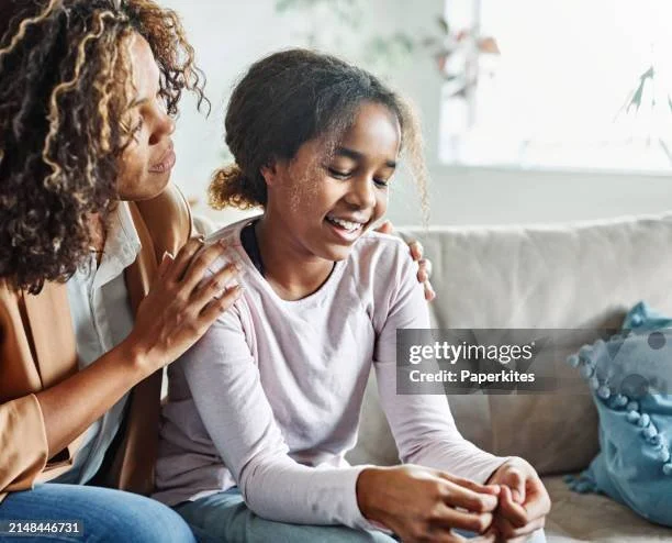 A girl sitting on a couch smiling, with a caregiver gently touching her shoulder